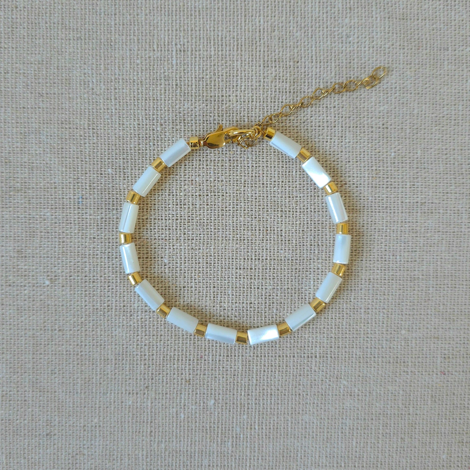 A delicate gold chain bracelet with alternating white rectangular beads and small gold spacer rings, displayed on a textured beige linen background.